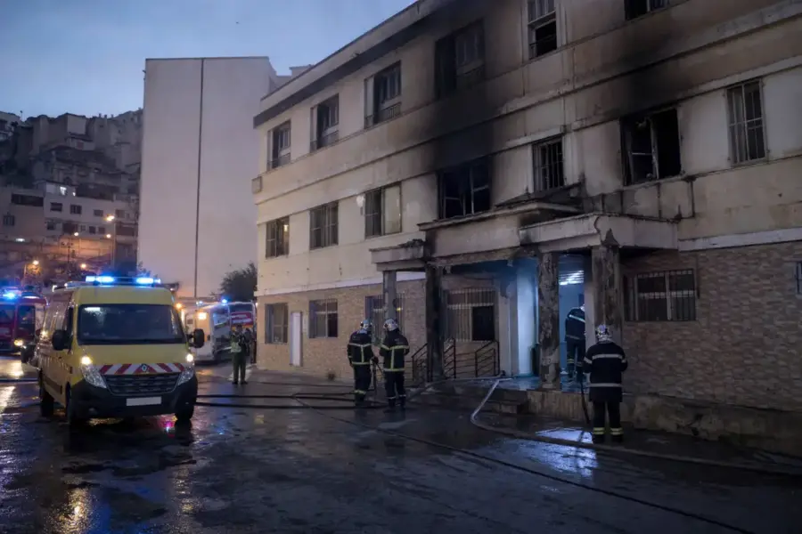 Vigili del fuoco e ambulanze davanti a un edificio annerito dal fumo dopo un incendio, con strada bagnata all’alba