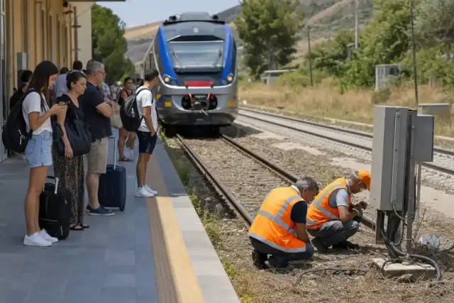 Passeggeri in attesa su un binario mentre due tecnici in giubbotti arancioni controllano un armadio cavi vicino ai binari