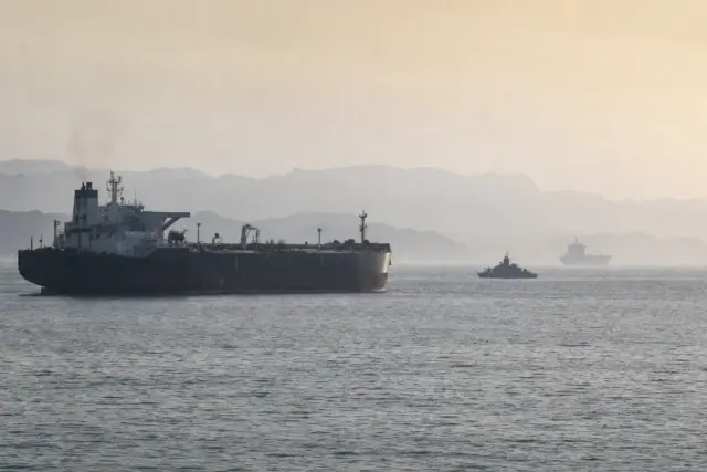 Petroliera in primo piano con piccola nave militare di pattugliamento e cargo lontano in mare tra foschia e montagne