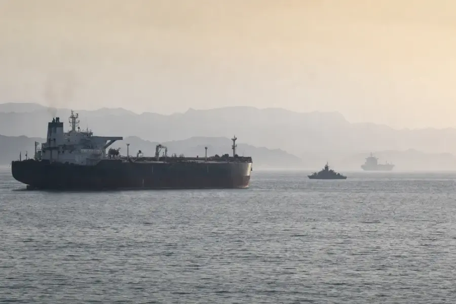 Petroliera in primo piano con piccola nave militare di pattugliamento e cargo lontano in mare tra foschia e montagne